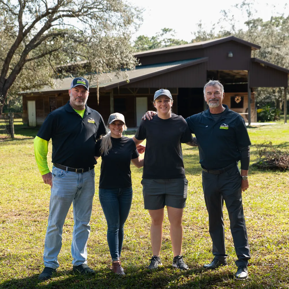 Four people standing arm in arm on a lawn in front of a barn, two of them in Gold Key Roofing shirts