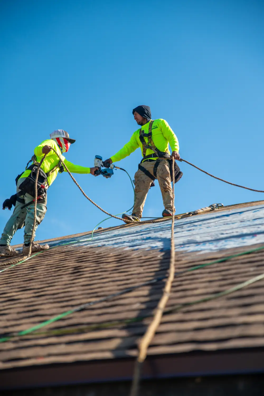 Two Gold Key roofers passing a nail gun on a half shingled roof against a blue sky