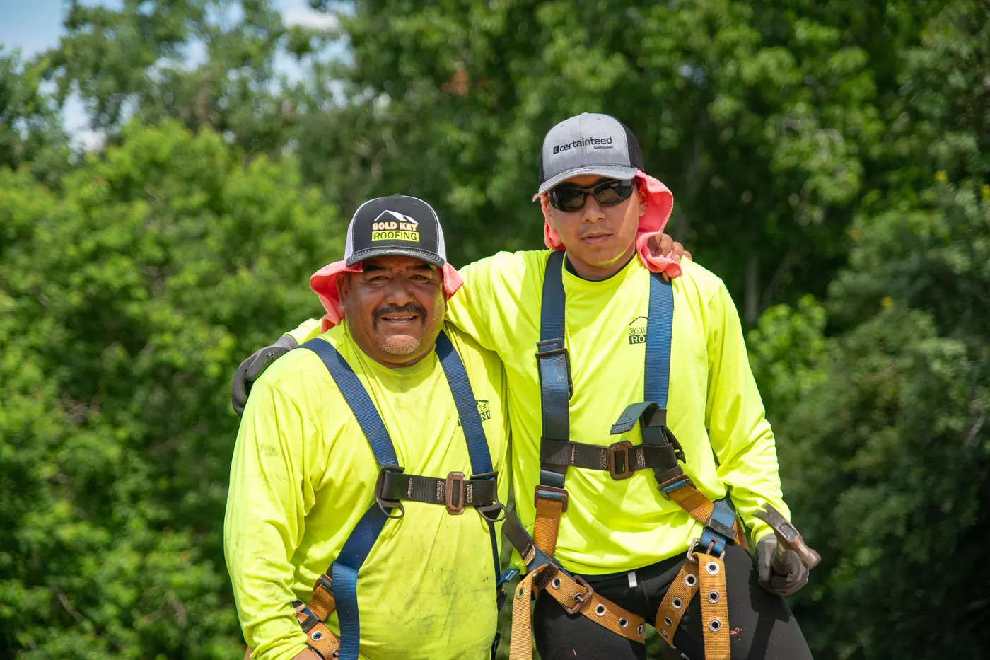 Two Gold Key Roofing roofers in safety harnesses smiling arm in arm on a roof